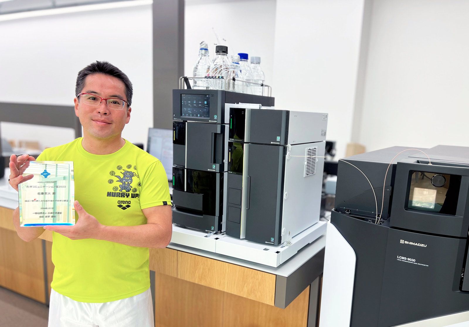 化学コミュニケーション賞受賞記念の盾を持つ佐藤先生。LC-MS分子構造解析装置の前で 化学コミュニケーション賞受賞記念の盾を持つ佐藤先生。LC-MS分子構造解析装置の前で